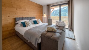 Un dormitorio con pared de madera, cama con cabecera gris, un banco con toallas y vistas a montañas a través de una ventana.