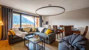 Una sala de estar moderna con sofá, sillón y una mesa de centro de vidrio. Se observa una mesa de comedor con sillas y vistas a montañas a través de amplios ventanales.