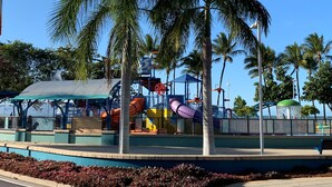 Un parque infantil con toboganes y estructuras para escalar, rodeado de palmeras y un cielo azul despejado.