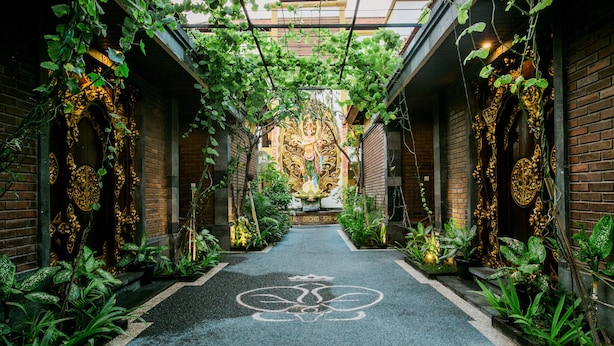 A narrow alleyway with brick walls and greenery, leading to a building with ornate decorations and a lotus symbol on the ground. - Royal Orchid Lodge (Nusa Dua)