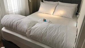 A neatly made bed with white linens and pillows, a bedside table with a lamp, and a window with sheer curtains. - Kalkedon Hostel (Istanbul)