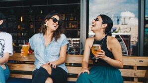 Three people sitting on a wooden bench at a bar with shelves of bottles and glasses in the background. - Kalkedon Hostel (Istanbul)