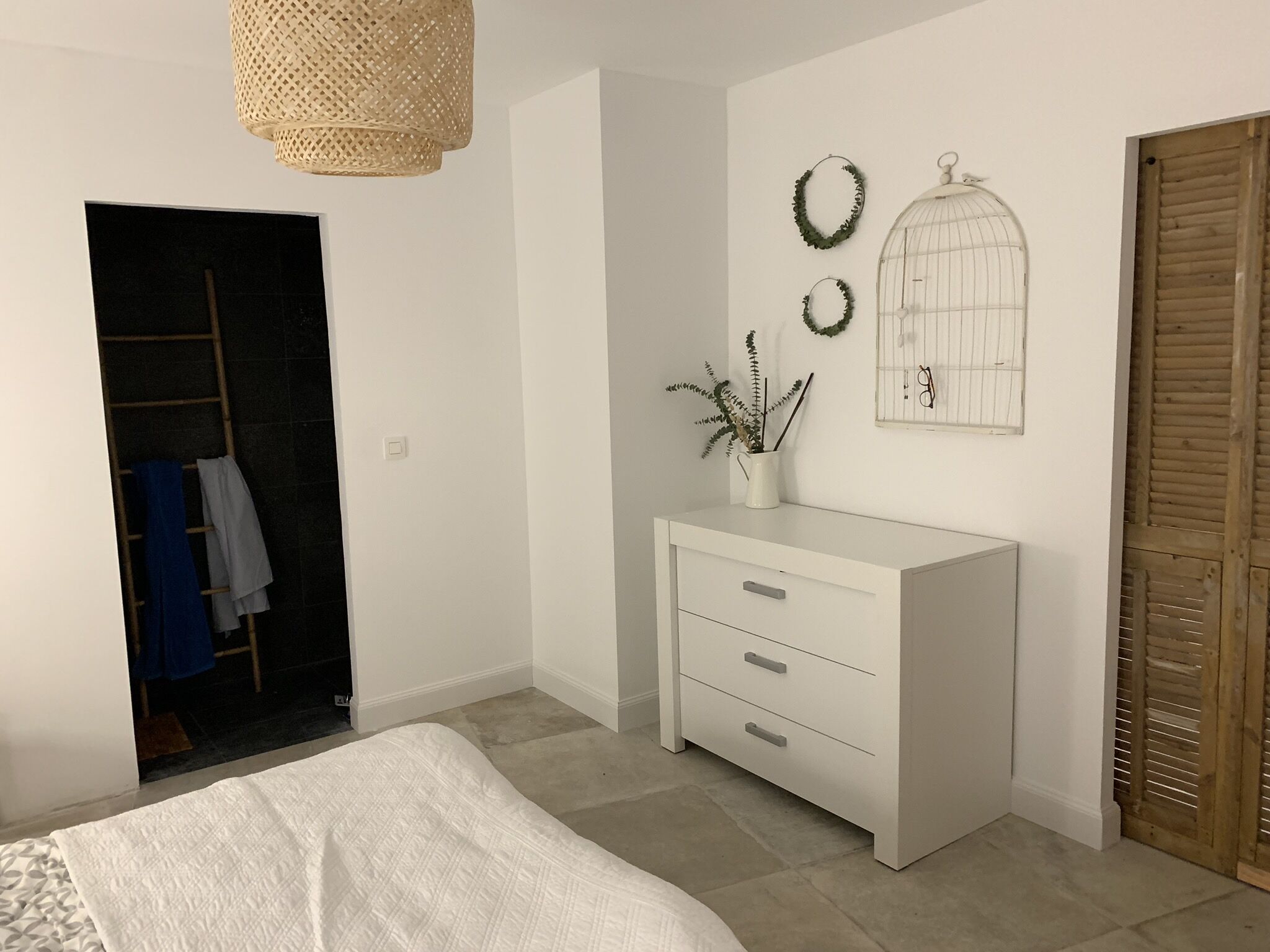 A bedroom with a bed, a white dresser, a wicker lamp, a wall-mounted birdcage, and a door with a wooden shutter.