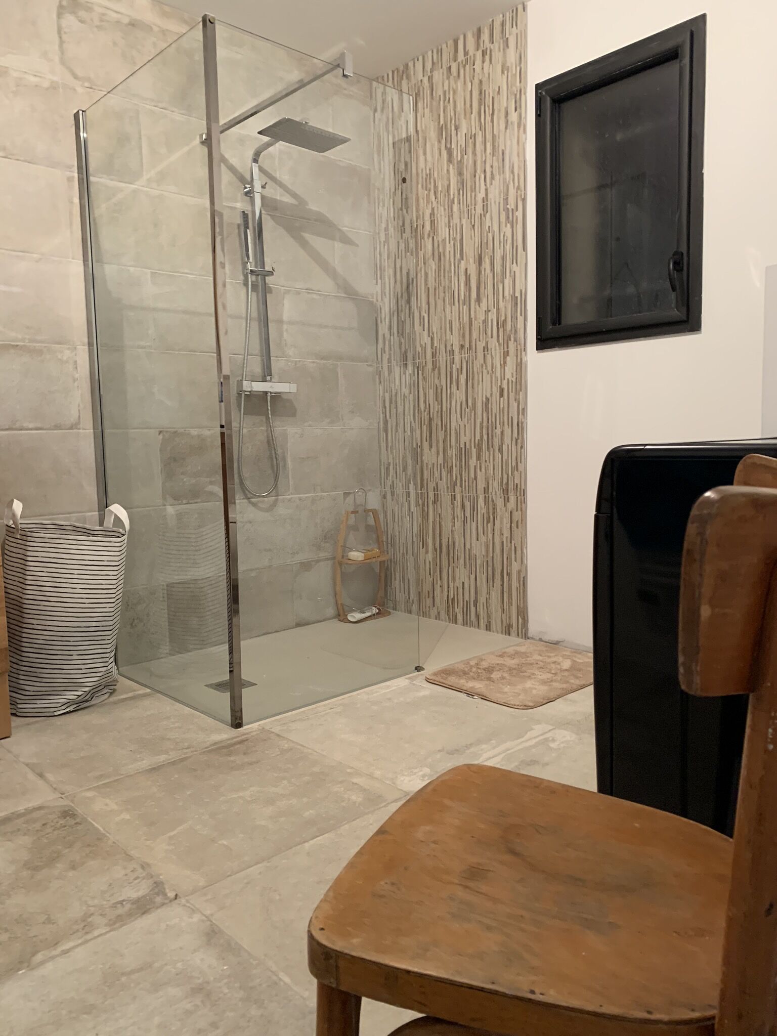 Un baño con cabina de ducha de vidrio, una silla de madera, una cesta de mimbre y un papel tapiz estampado.
