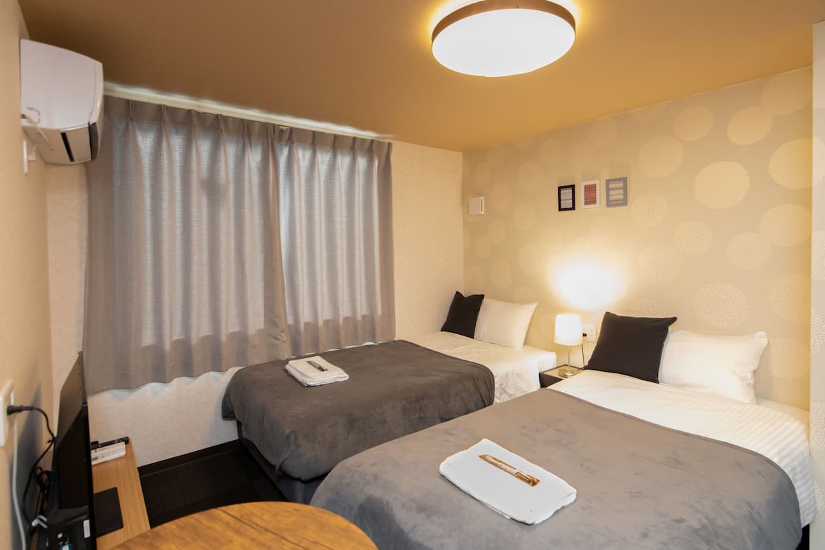 Quarto de hotel com duas camas, uma escrivaninha e uma cadeira. Há uma janela com cortinas, ar-condicionado fixado na parede e iluminação no teto.