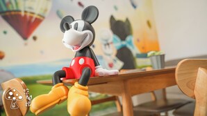 A Mickey Mouse figurine seated at a wooden table with a blurred background featuring a cartoon character and a hot air balloon. - Youge Homestay (Taitung)