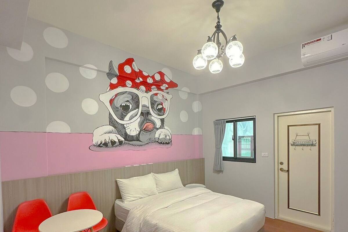 A bedroom with a bed, a red chair, a small table, a ceiling light, and a wall with a polka dot pattern.
