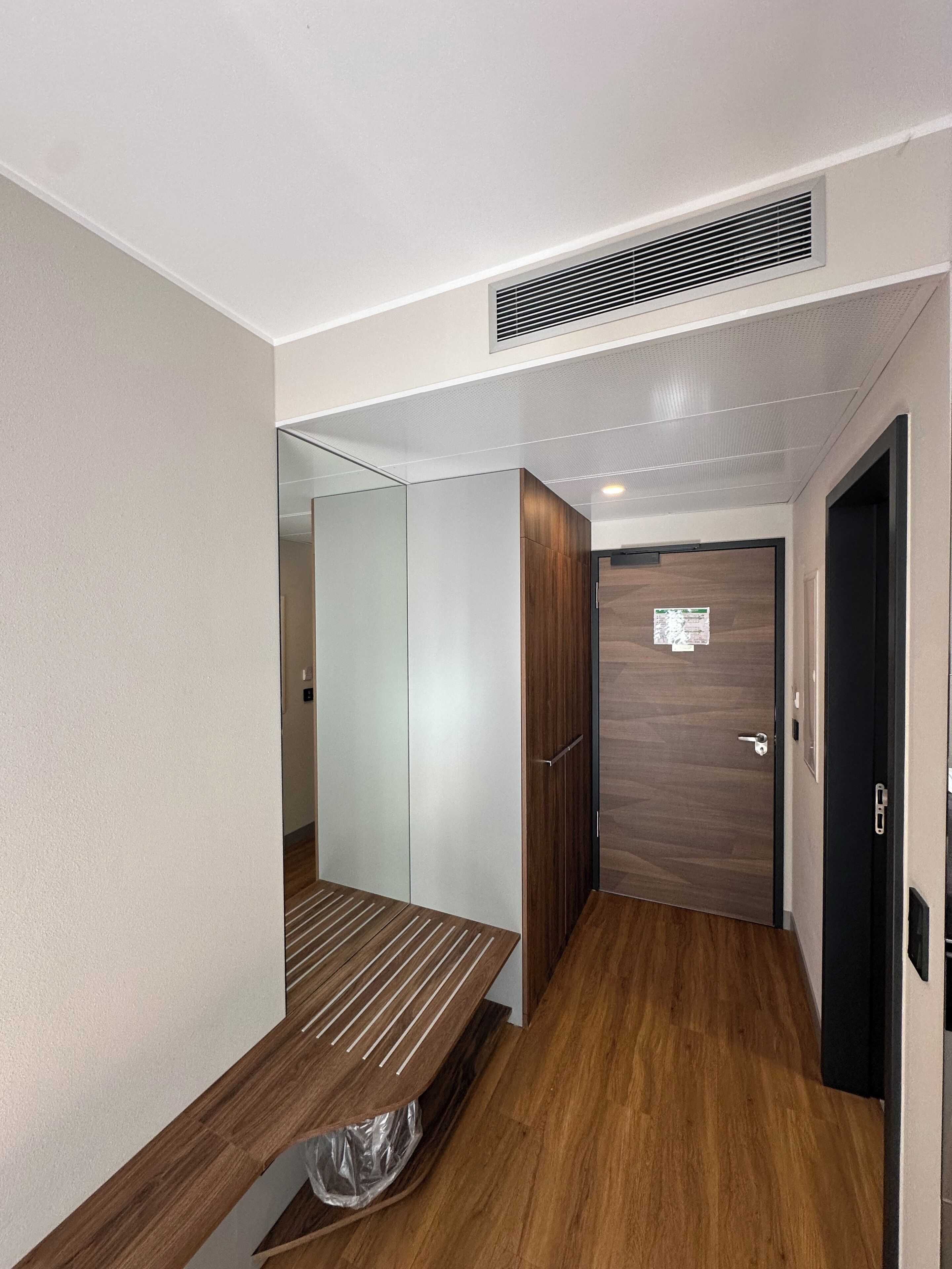 Un pasillo estrecho con un banco de madera, una puerta con un cartel y una rejilla de ventilación en el techo.