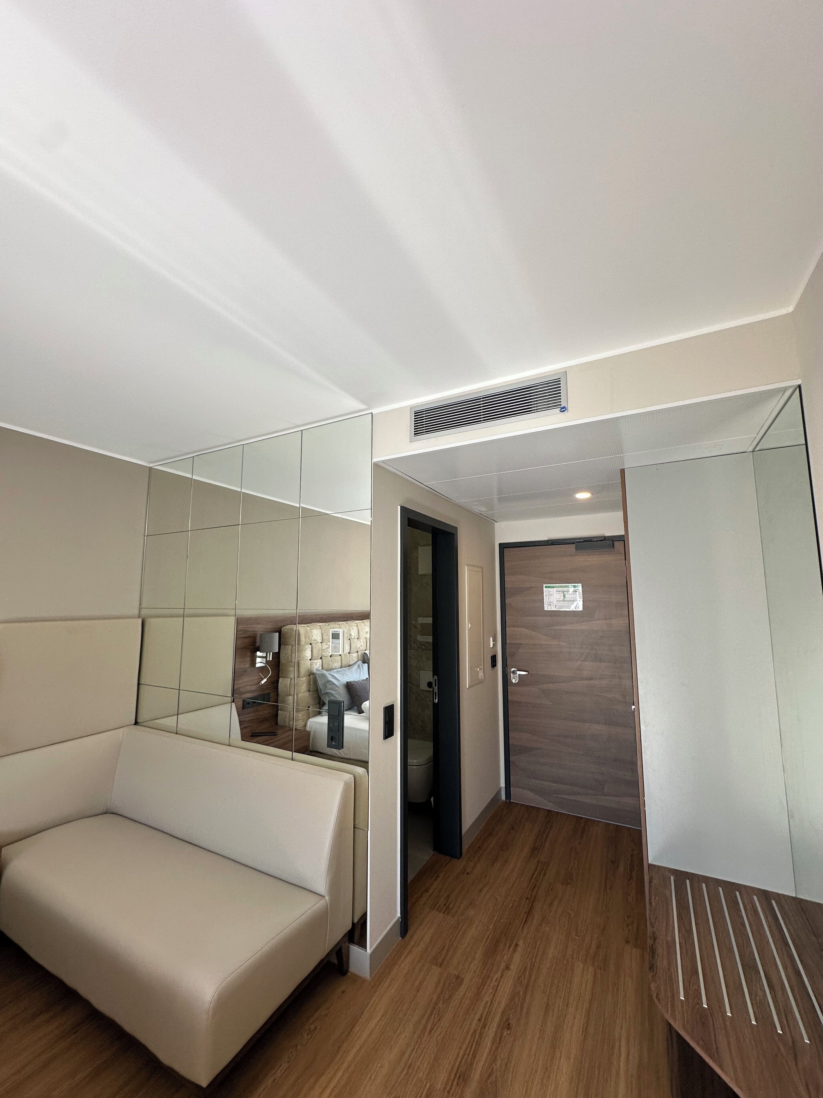 A modern hotel room with a sofa, a wooden floor, a mirror, and a door.