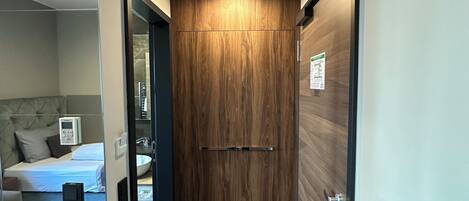 Un pasillo de hotel con una puerta de madera, una cama con cabecera, una mesita de noche y un panel de control en la pared.
