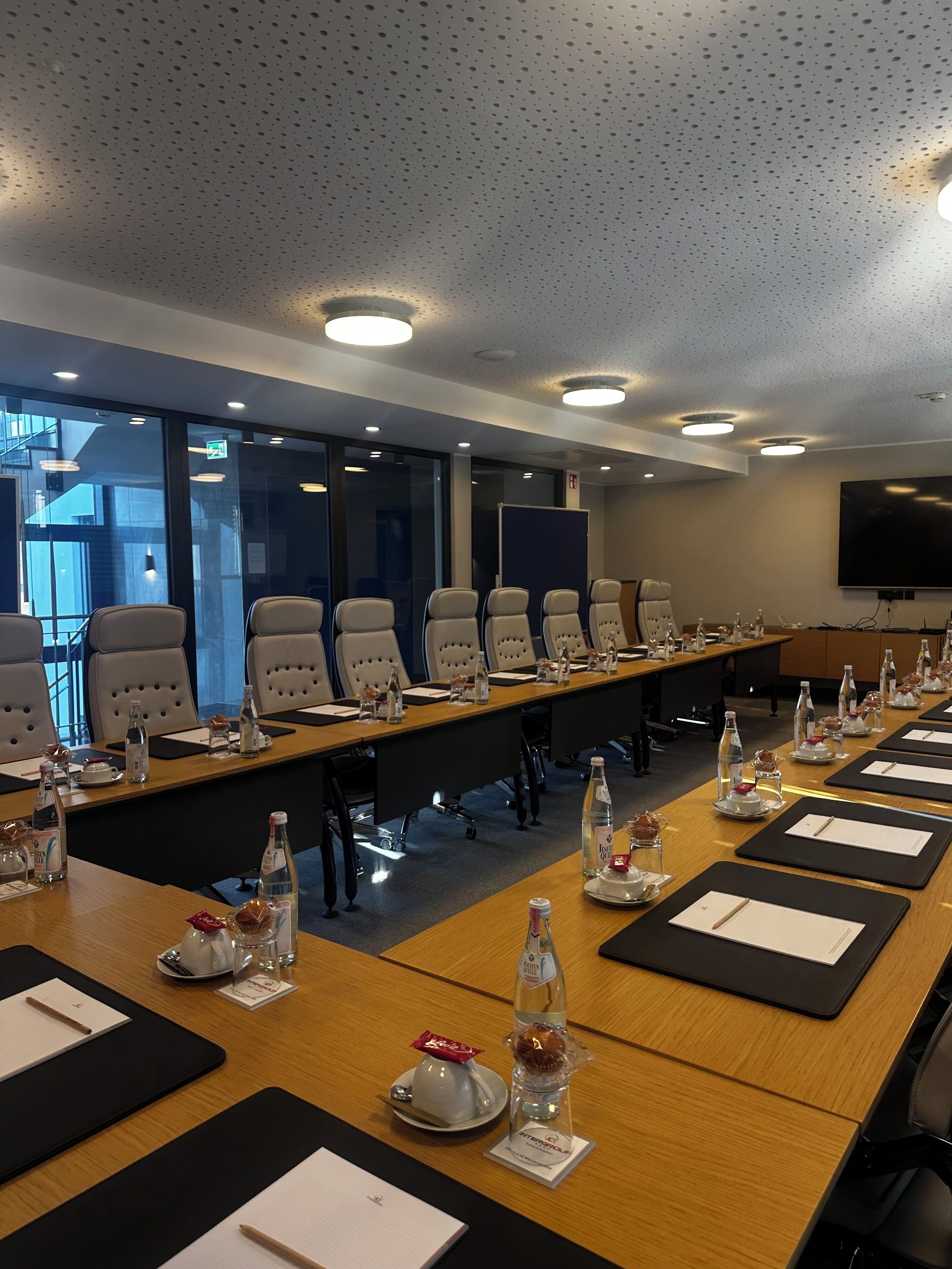 Un salón de conferencias con una larga mesa, sillas, tarjetas de identificación, botellas de agua y una pantalla de televisión.