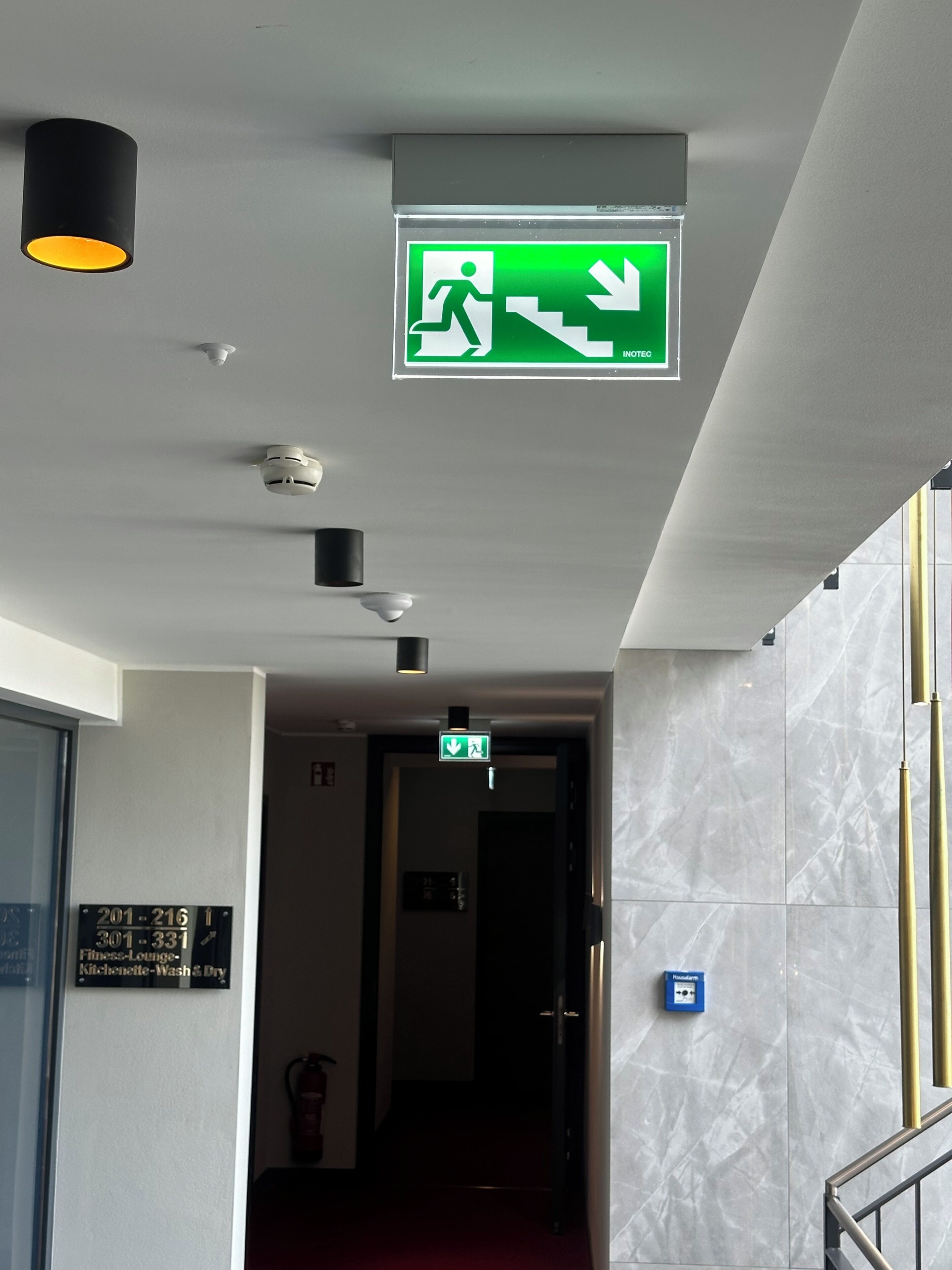 Pasillo de hotel con señal de salida de emergencia, detector de incendios y letrero que indica el gimnasio, kitchenette y lavandería.