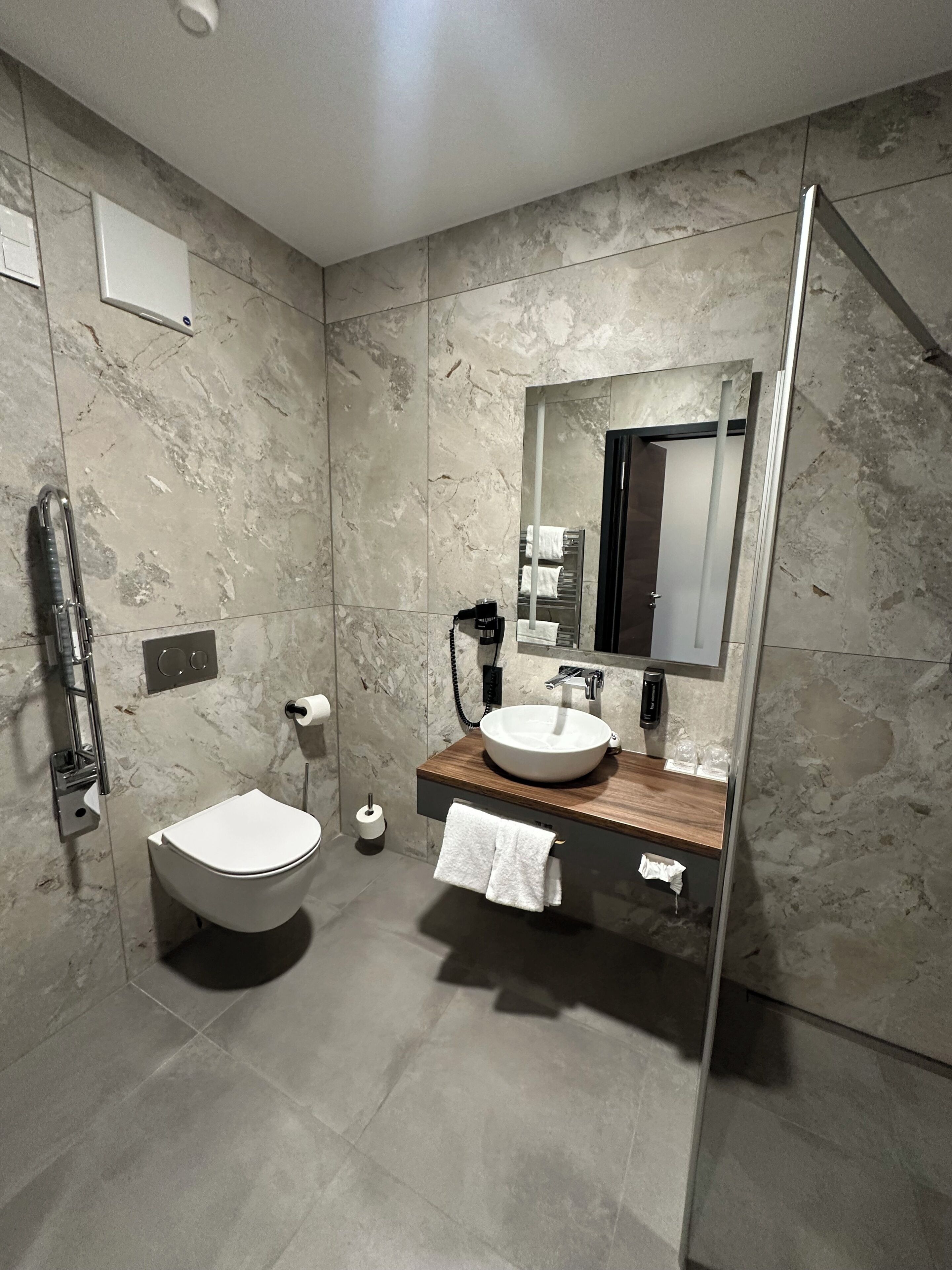 Une salle de bain moderne avec un mur en marbre, des toilettes blanches, un meuble-lavabo en bois doté d’un grand miroir et une cabine de douche en verre.