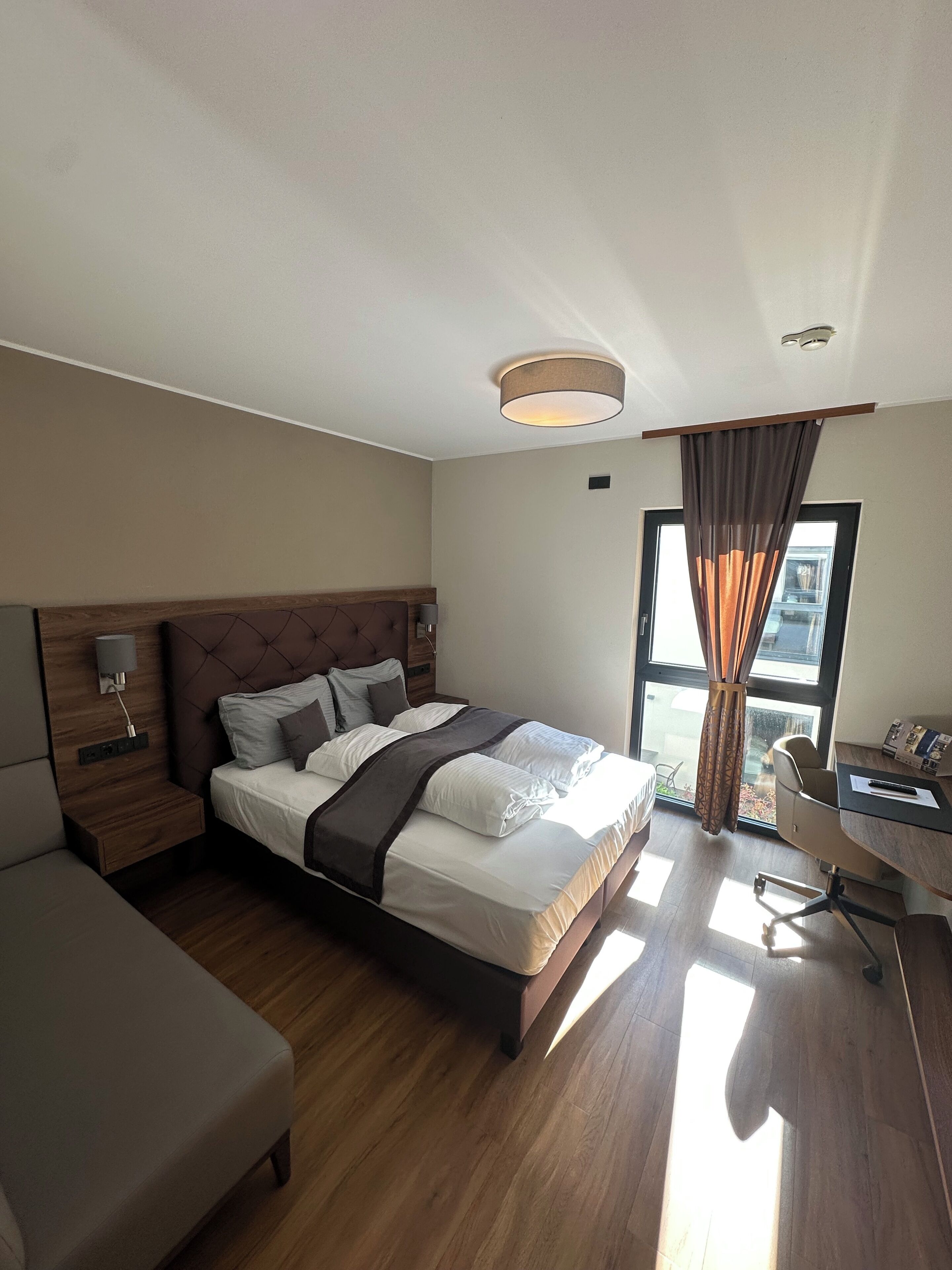 A hotel room with a large bed, a desk, a chair, a window with curtains, and a ceiling light.