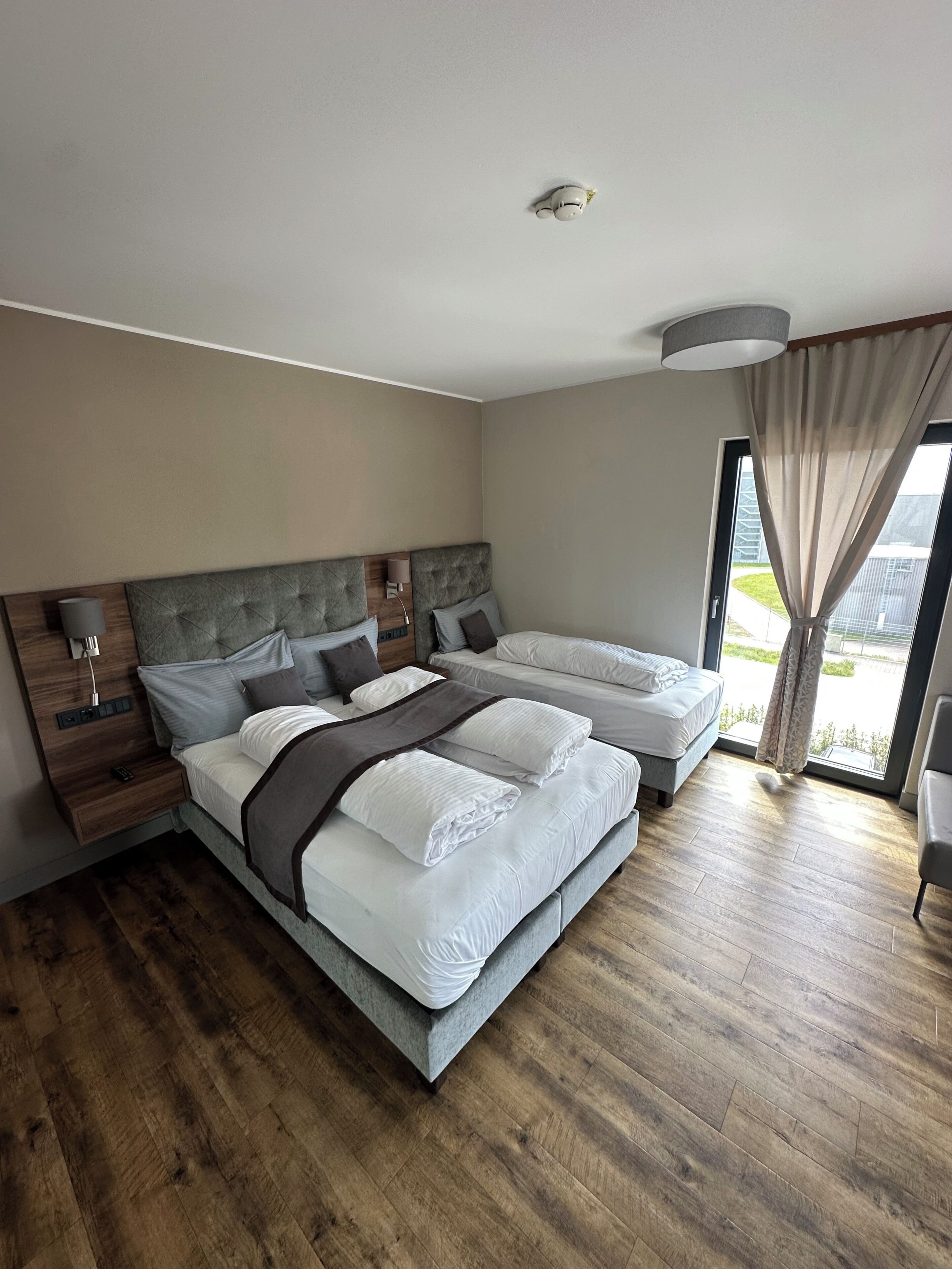 A modern bedroom with a large bed, bedside tables, a lamp, and a view through a window.