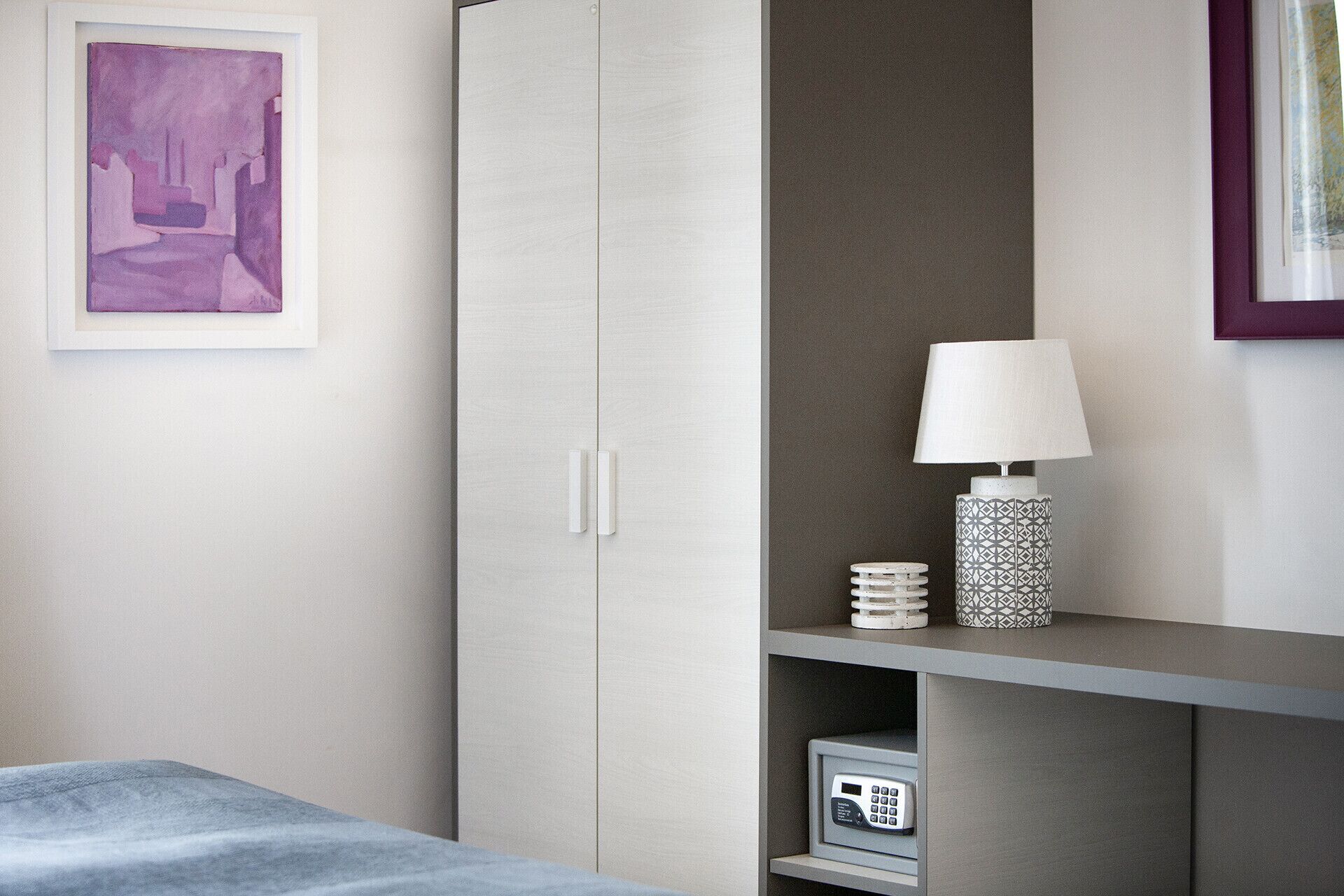 A bedroom with a bed, a wardrobe, a shelf with a lamp, and a framed picture on the wall.