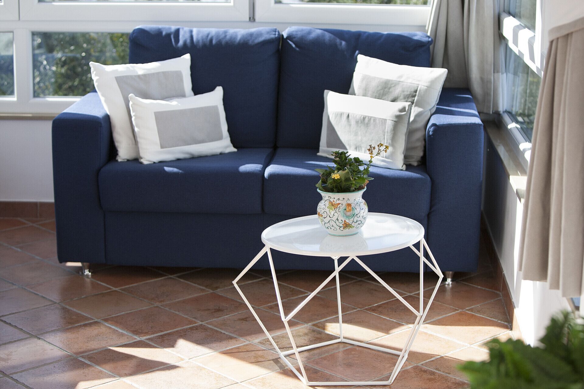 Ein blaues Sofa mit weißen Kissen, ein weißer Tisch mit geometrischem Fuß und eine Topfpflanze.