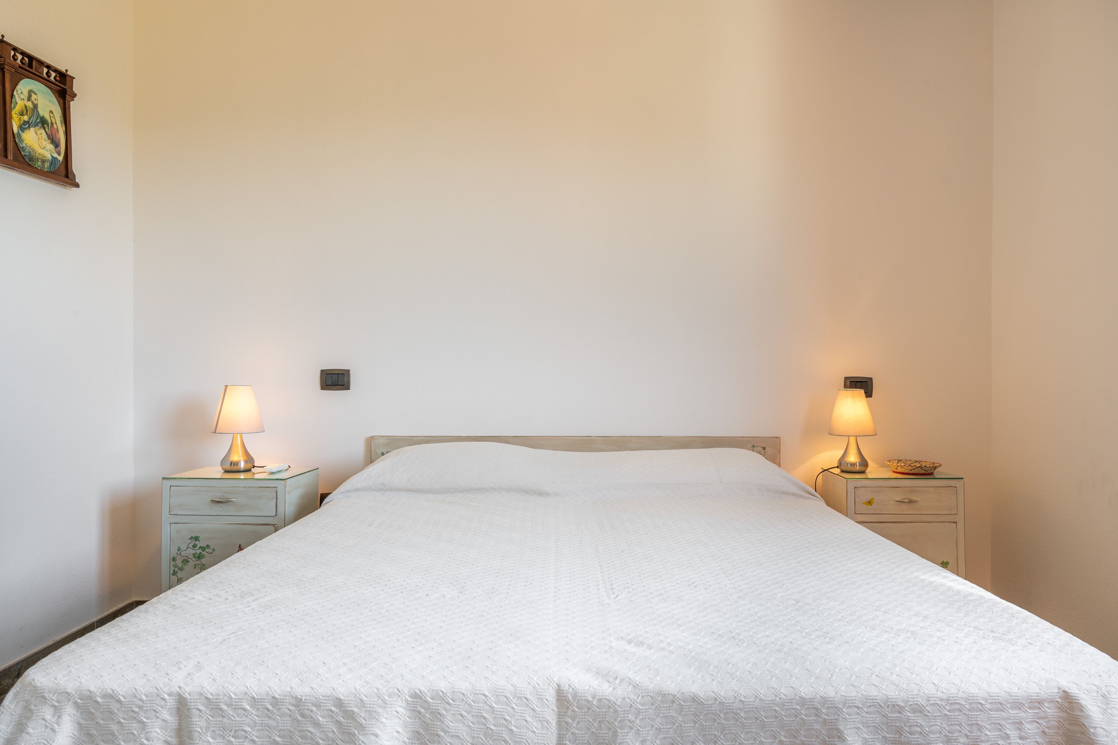 Een eenpersoonsbed met een witte dekbedovertrek in een kamer met effen muren. Aan beide zijden van het bed staan twee nachtkastjes met lampen.