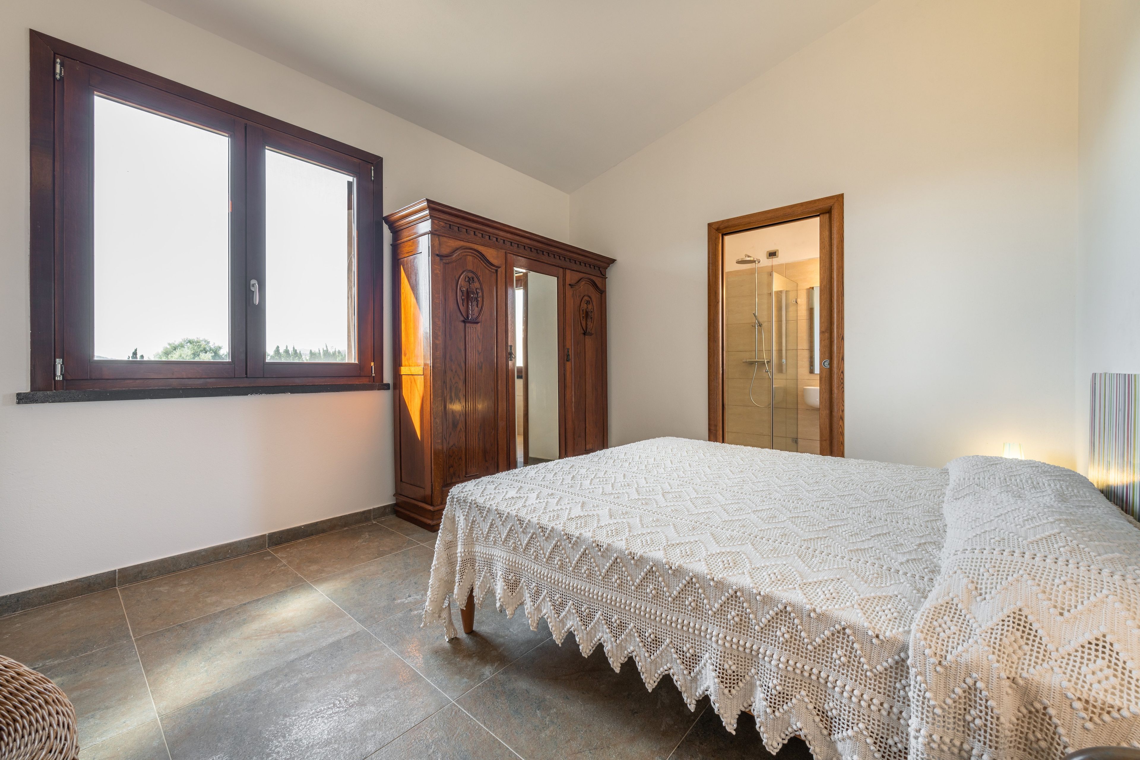 A bedroom with a large window, a wardrobe, a bed with a white bedspread, and a shower area.