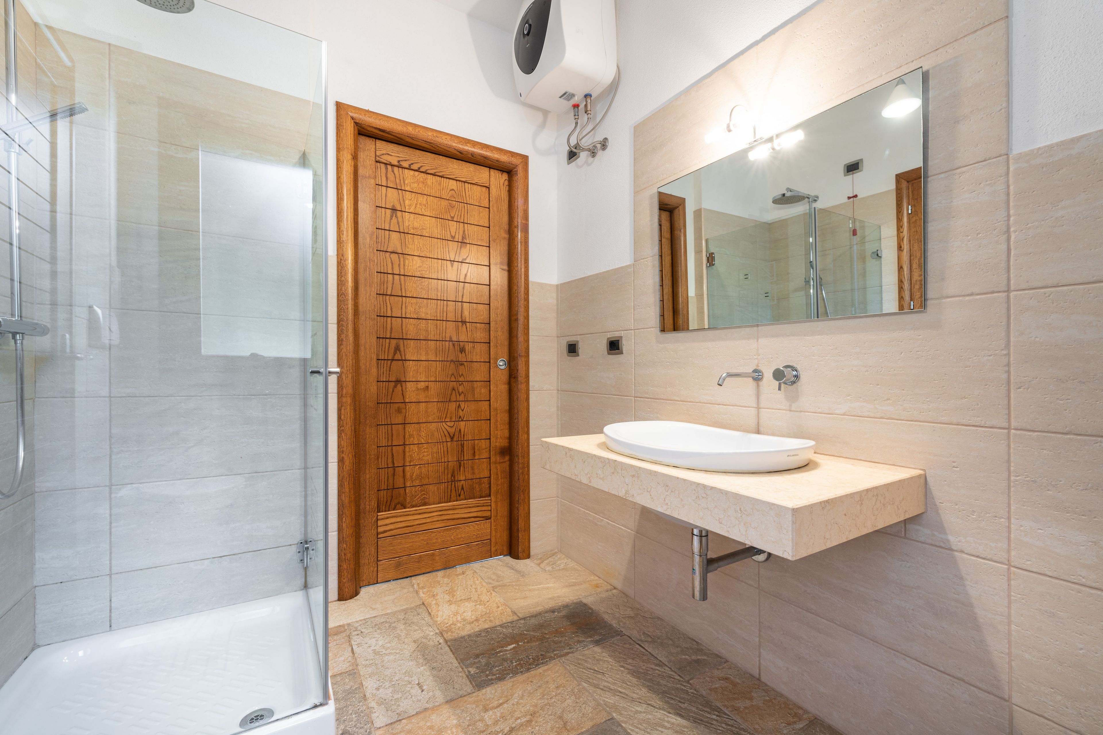 Un baño moderno con cabina de ducha de vidrio, puerta de madera, lavamanos blanco y un espejo grande.