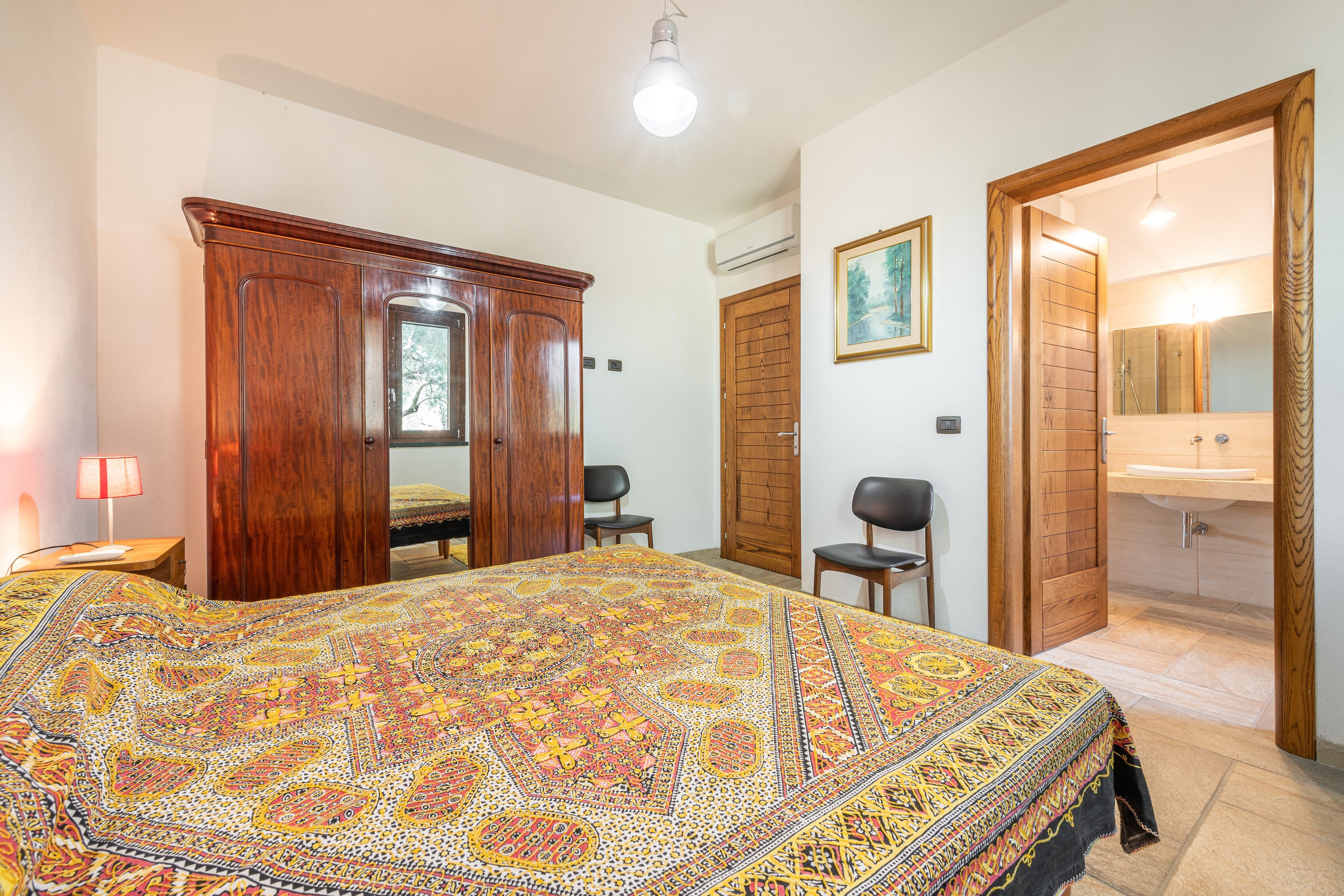 Un dormitorio con una cama grande, un armario, una silla, un escritorio y un baño con lavabo y espejo.