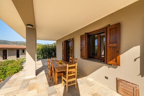 Eine überdachte Terrasse mit Holztisch und -stühlen, Blick auf die umliegende Landschaft und ein Gebäude mit Ziegeldach im Hintergrund.