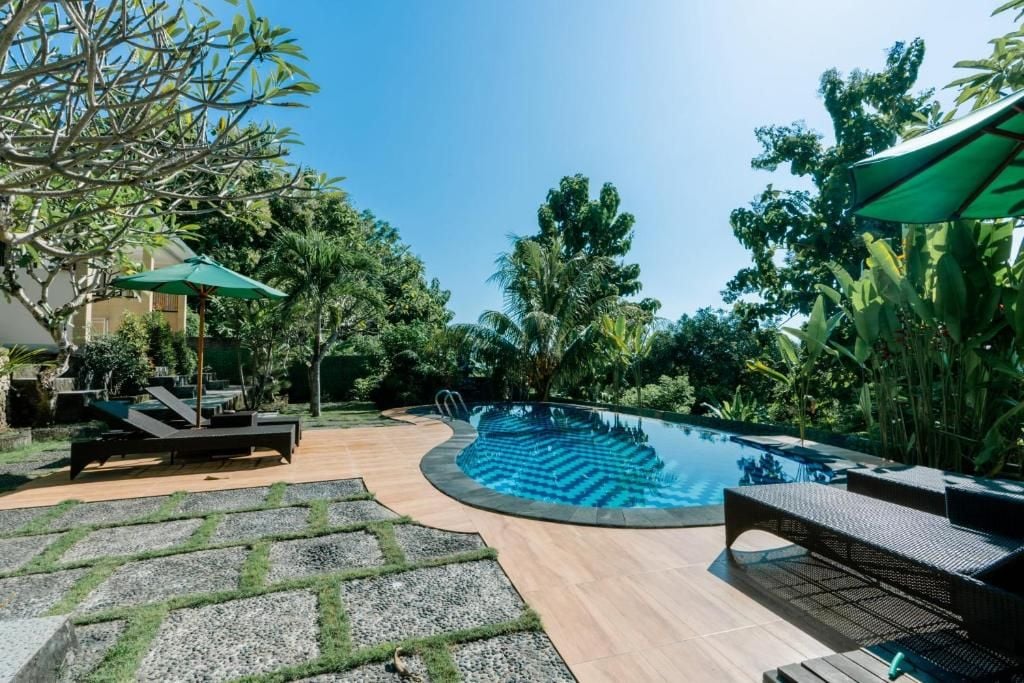Una piscina a forma sinuosa circondata da poltrone a sdraio e ombrelloni, immersa in un giardino curato con alberi e cielo sereno.