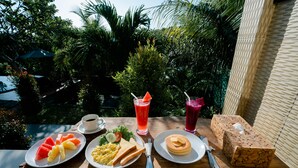 Mesa de madeira com itens de café da manhã, incluindo melancia, torradas, ovos, panquecas e bebidas, com um fundo de vegetação exuberante.