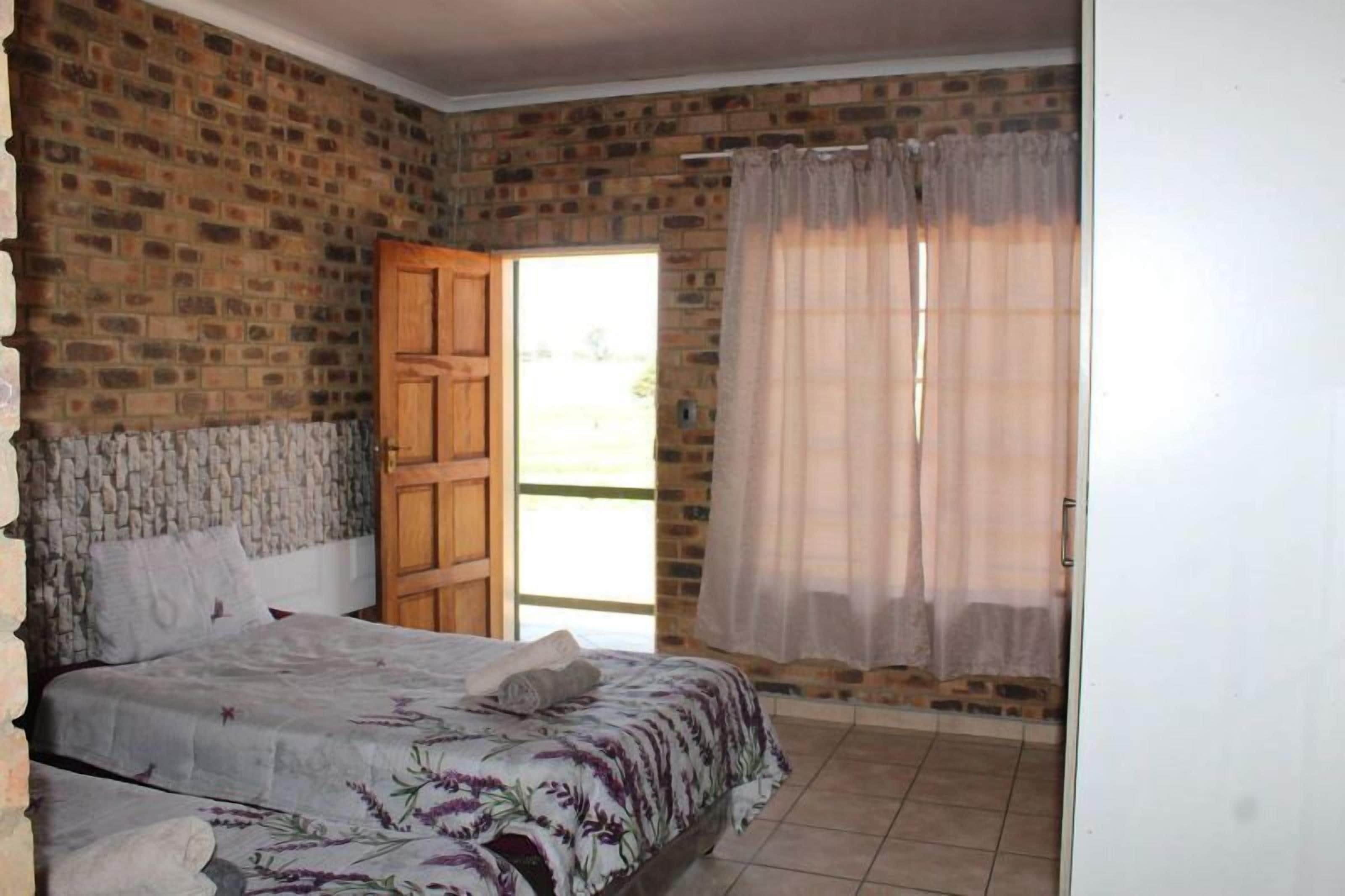 A bedroom with a brick wall, a wooden door, a bed with a floral bedspread, and a window with sheer curtains.