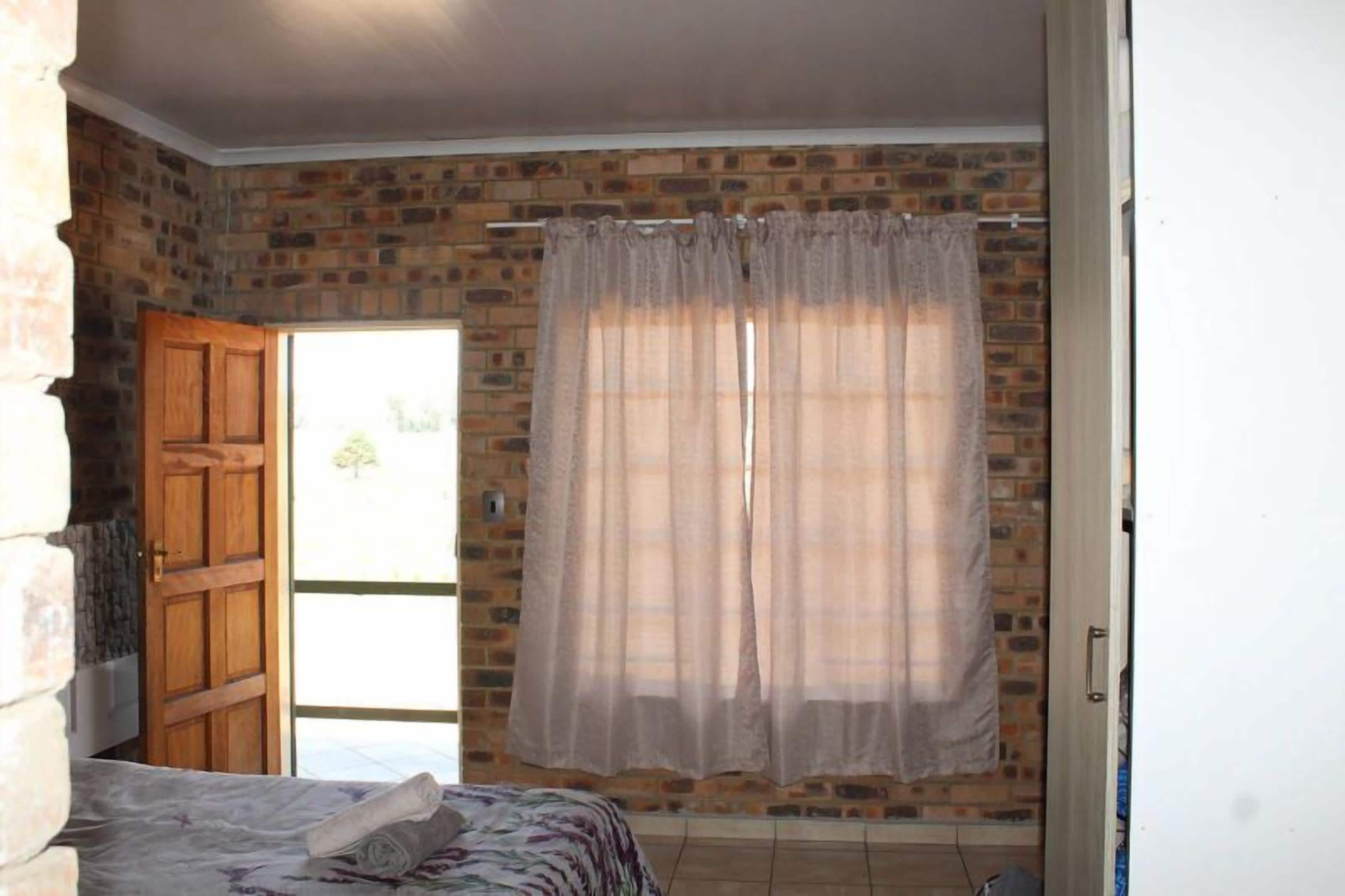 Una habitación con pared de ladrillo, puerta de madera, cama con cubrecama estampado y ventana con cortinas traslúcidas.