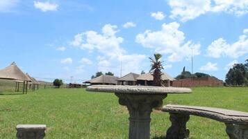 Un campo cubierto de hierba con un banco de piedra y una mesa de piedra en primer plano, y un edificio con techo de paja al fondo.