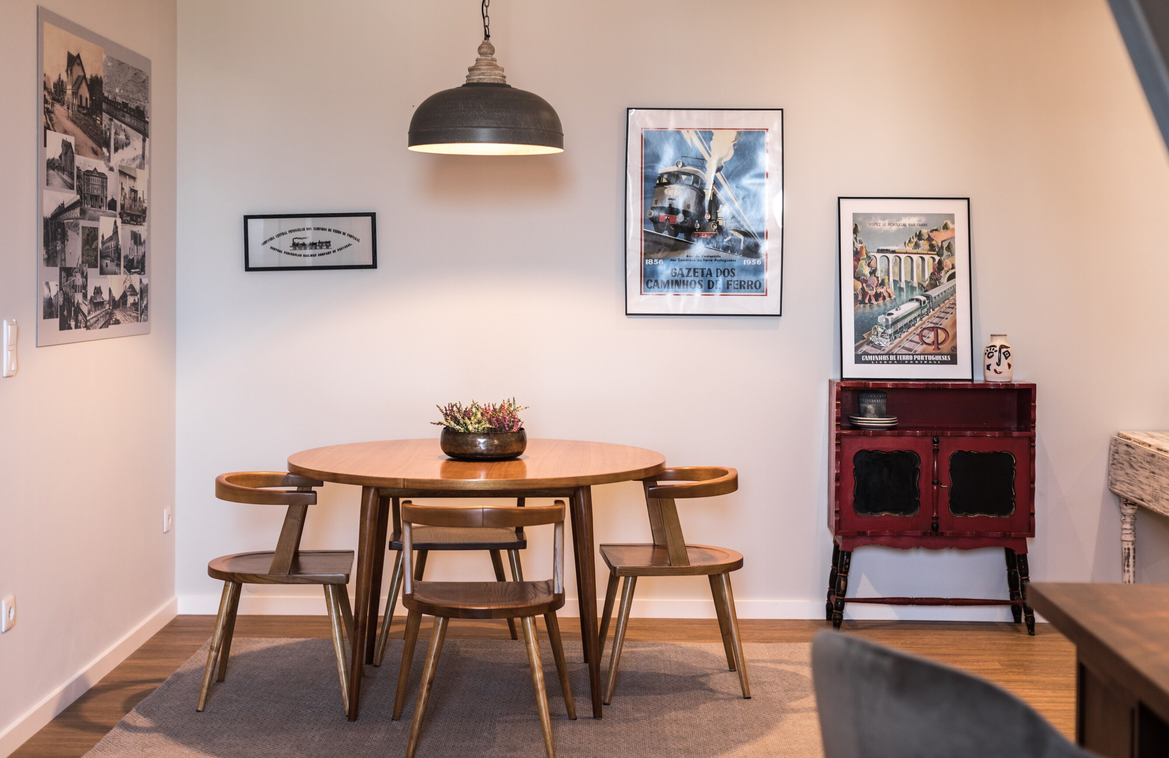 A dining area with a round wooden table and four chairs, a hanging light fixture, and three framed posters on the wall.