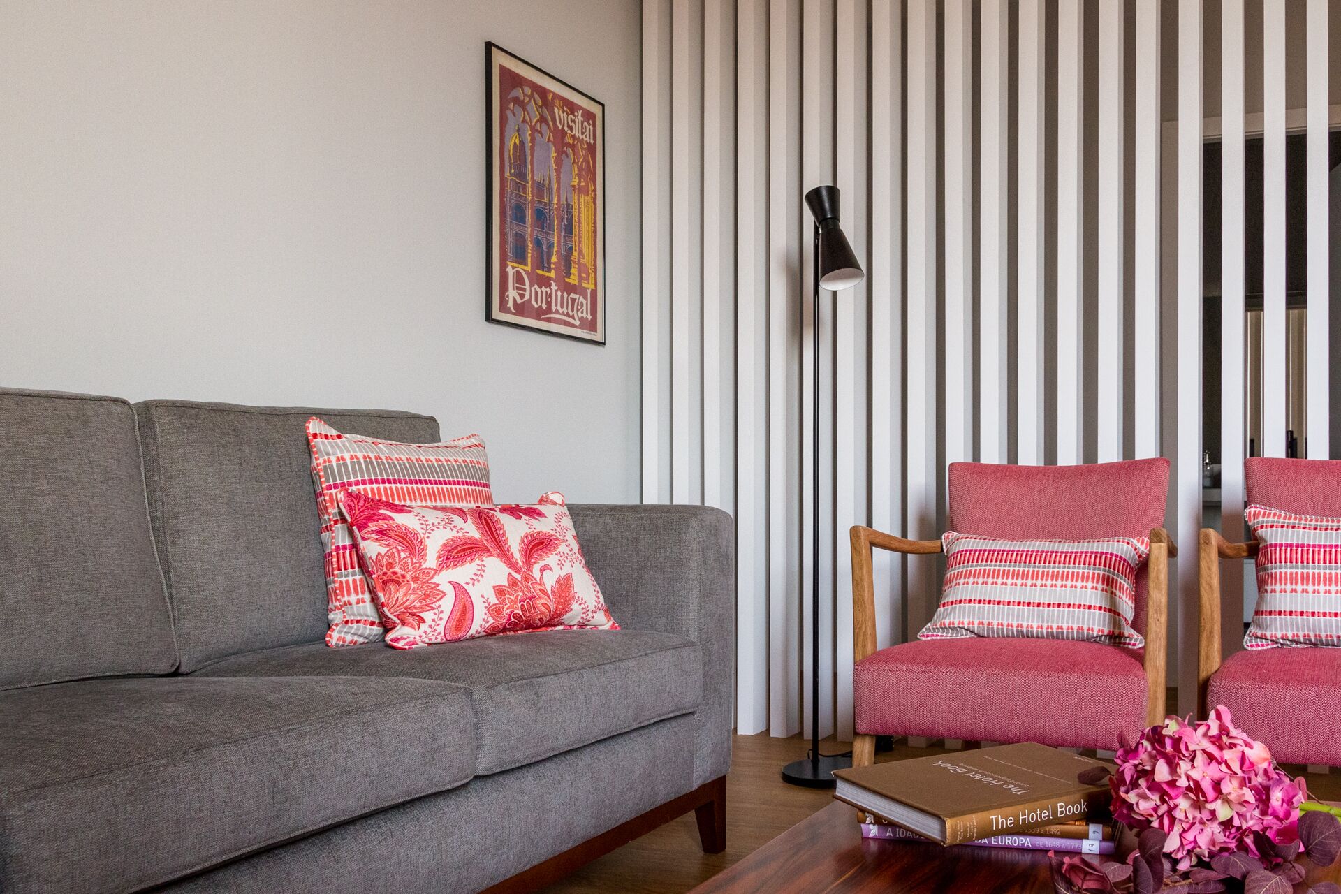 A living room with a grey sofa, red and white patterned cushions, a wooden chair with similar cushions, a floor lamp, and a framed picture on the wall.