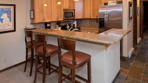 Una cocina con armarios de madera, un refrigerador de acero inoxidable, un microondas y una encimera de granito. Hay dos taburetes de bar de madera y luces colgantes sobre la encimera.