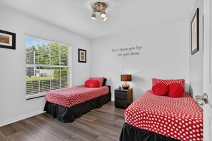A bedroom with two beds, a window with blinds, a bedside table with a lamp, and a motivational quote on the wall.