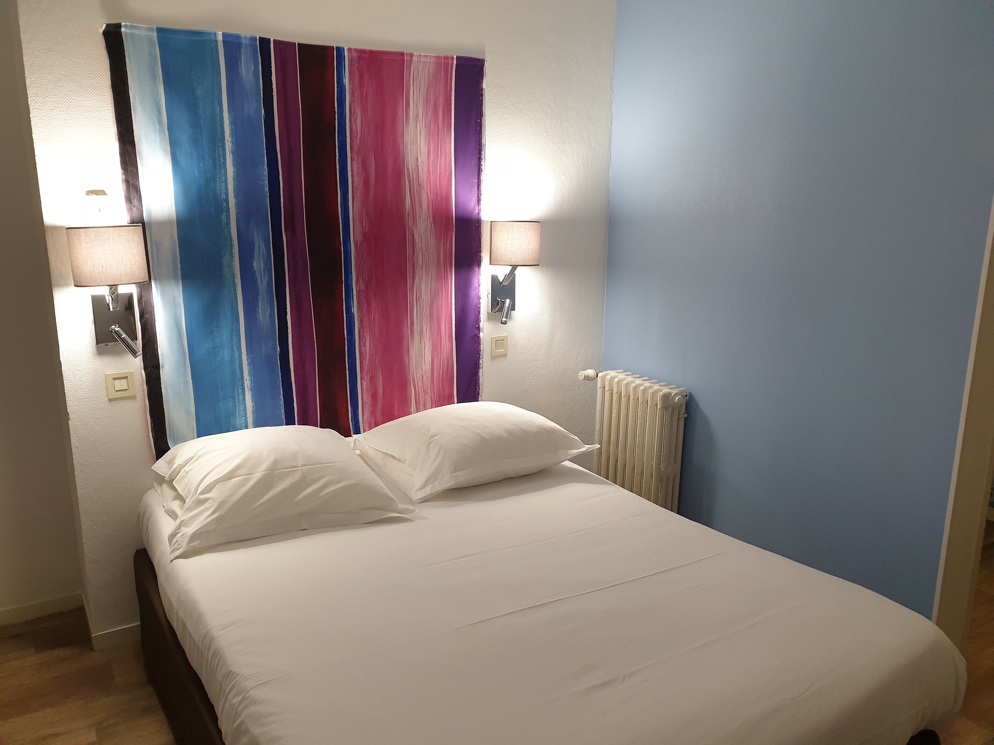 A hotel room with a bed, two wall-mounted lamps, a radiator, and a colorful striped curtain.