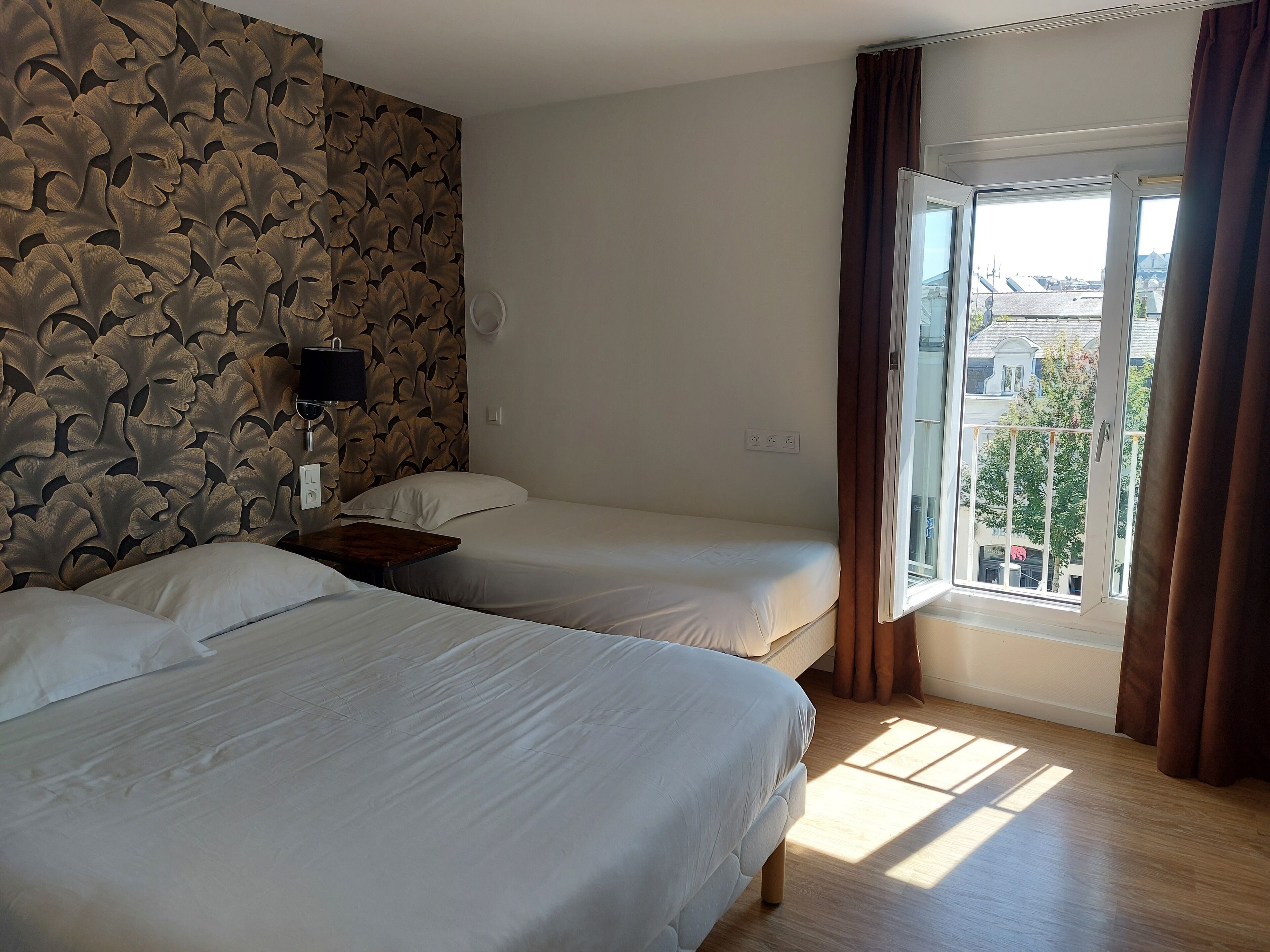 A hotel room with two beds, a window with curtains, a wall with a leaf pattern, and a view of buildings outside.