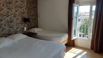 A hotel room with two beds, a window with curtains, a wall with a leaf pattern, and a view of buildings outside.