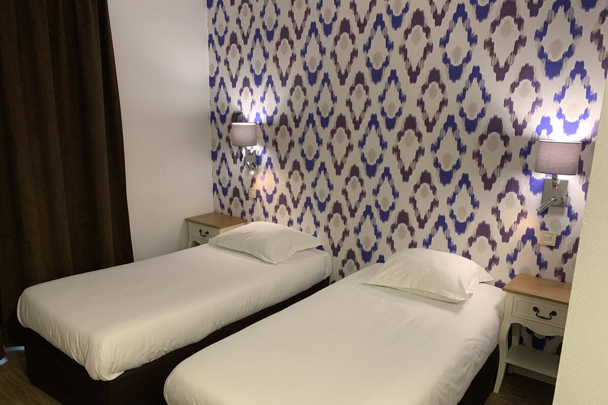 A hotel room with two single beds, a patterned wall, bedside lamps, and a wooden floor.