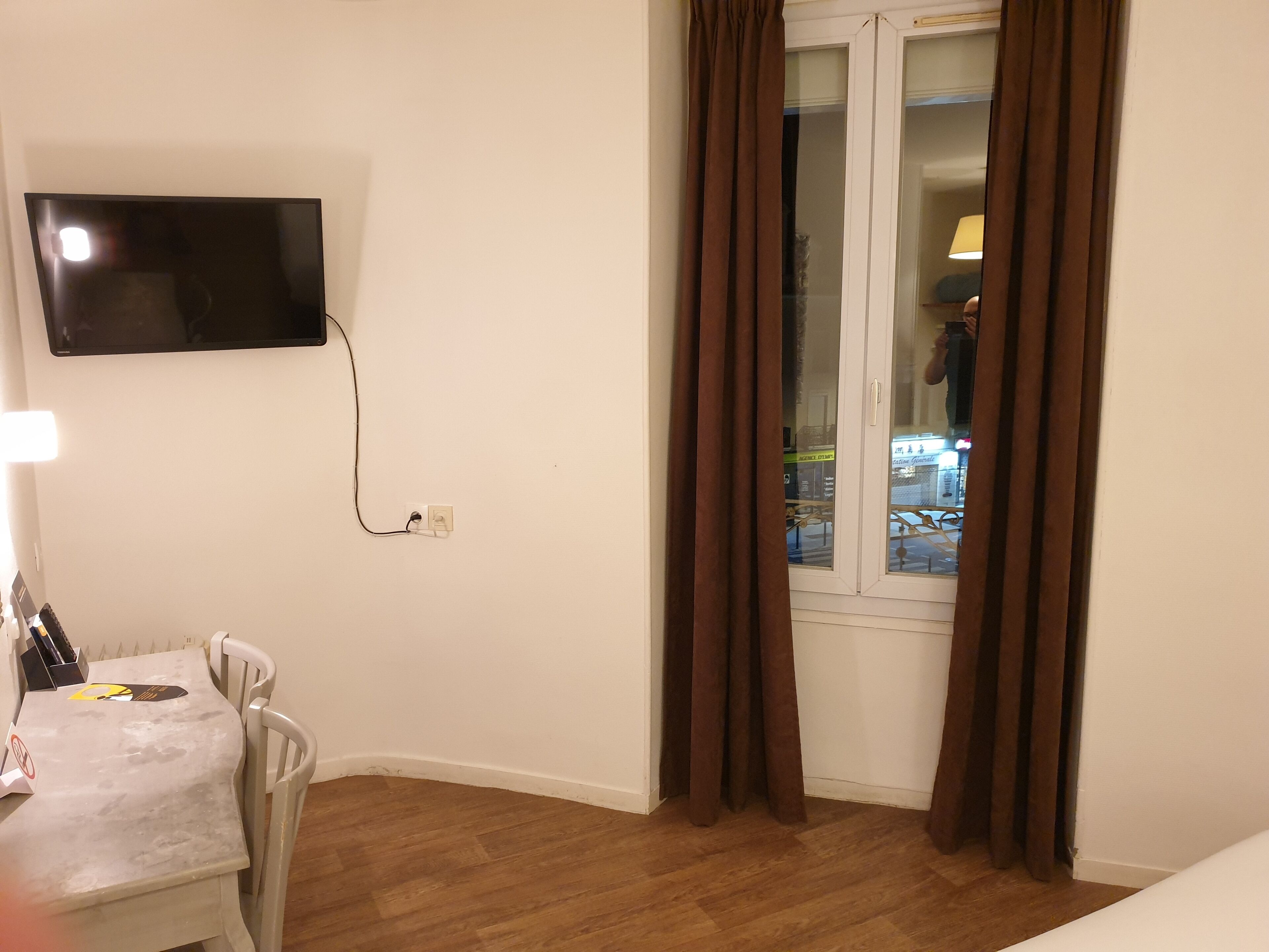 A hotel room with a flat-screen TV mounted on the wall, a desk with a chair, and a window with curtains.