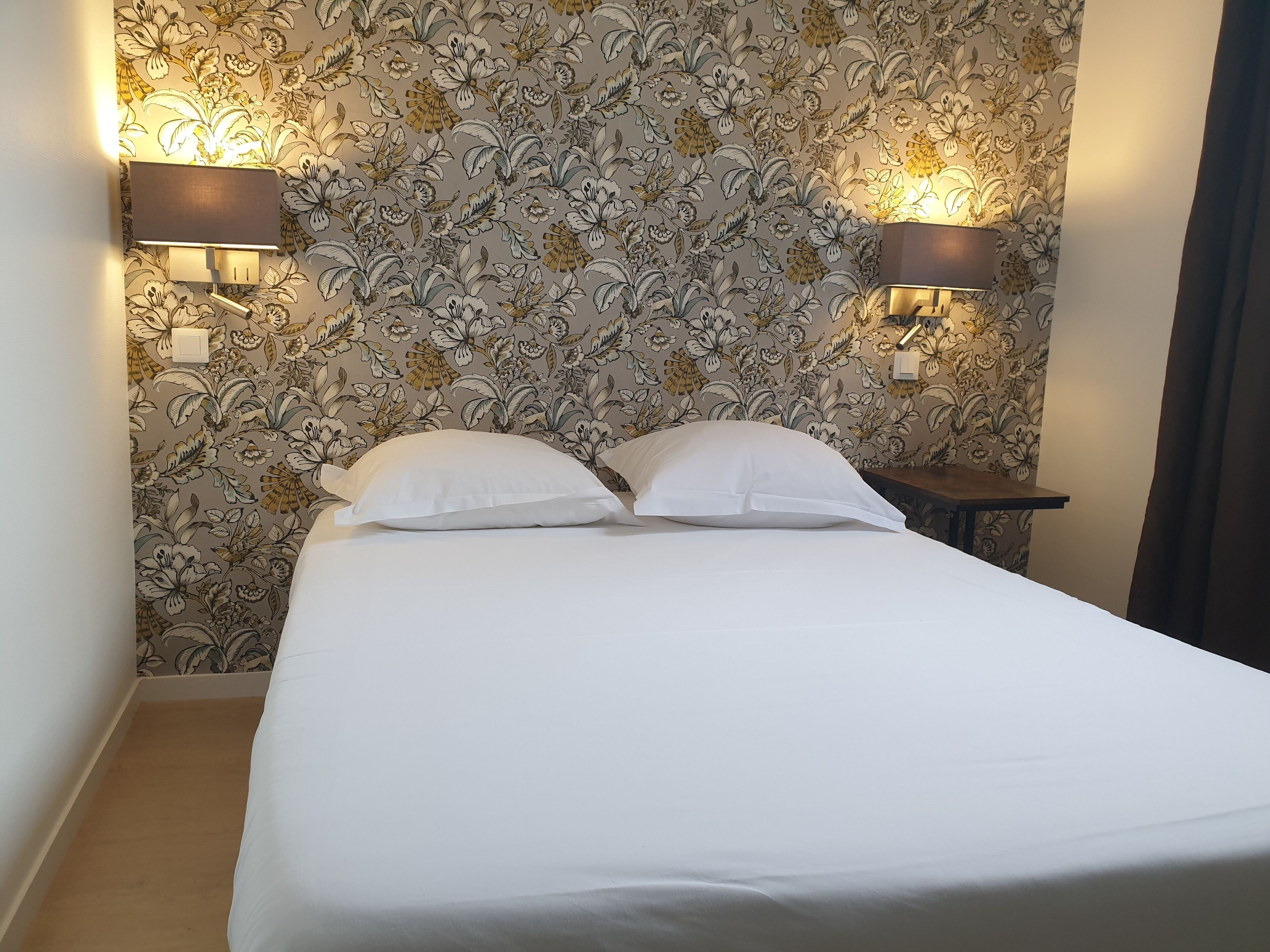 A neatly made bed with white linens in a room with floral wallpaper and two wall-mounted lamps.