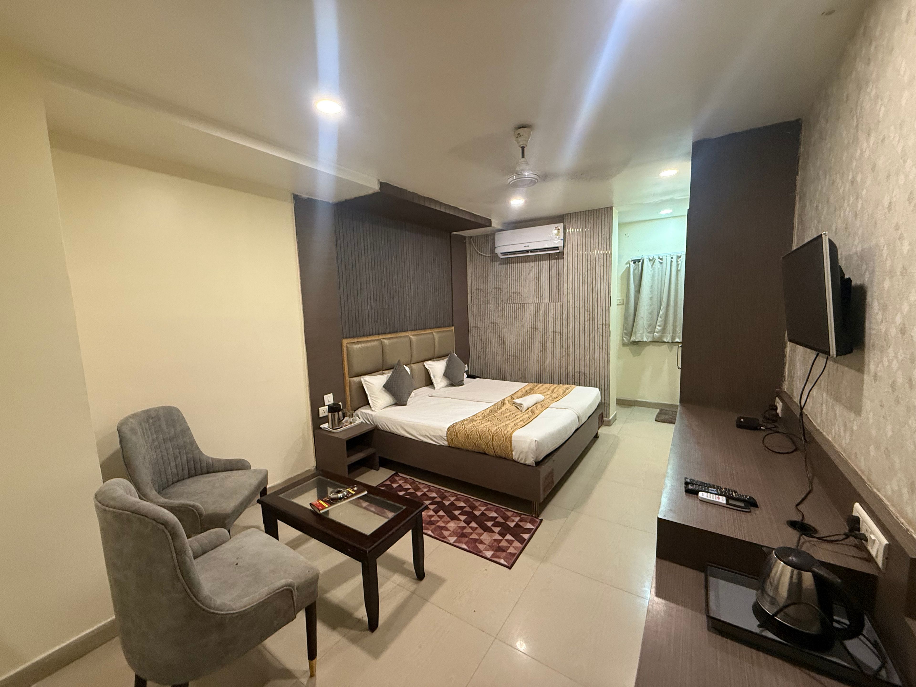 A hotel room with a bed, a seating area with a chair and a table, a desk with a computer, and a television mounted on the wall.