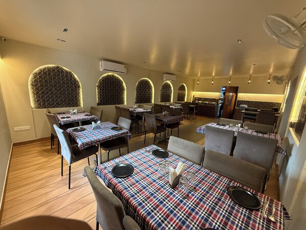 A restaurant with tables covered in plaid tablecloths, arched windows, and a kitchen area in the background. - Hotel Pratap Residency (Ranchi)