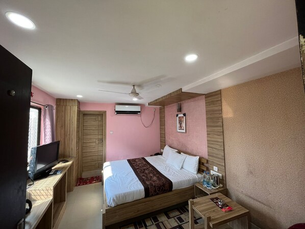 A hotel room with a bed, a TV, a ceiling fan, an air conditioning unit, and a small table with a bottle and a remote control. - Hotel Pratap Residency (Ranchi)