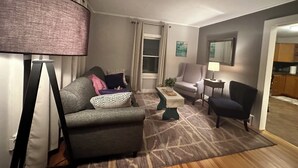 A modern living room with a grey sofa, a small side table, a floor lamp, and a view into the kitchen. - Nestled in the Beekman Art District (Saratoga Springs)
