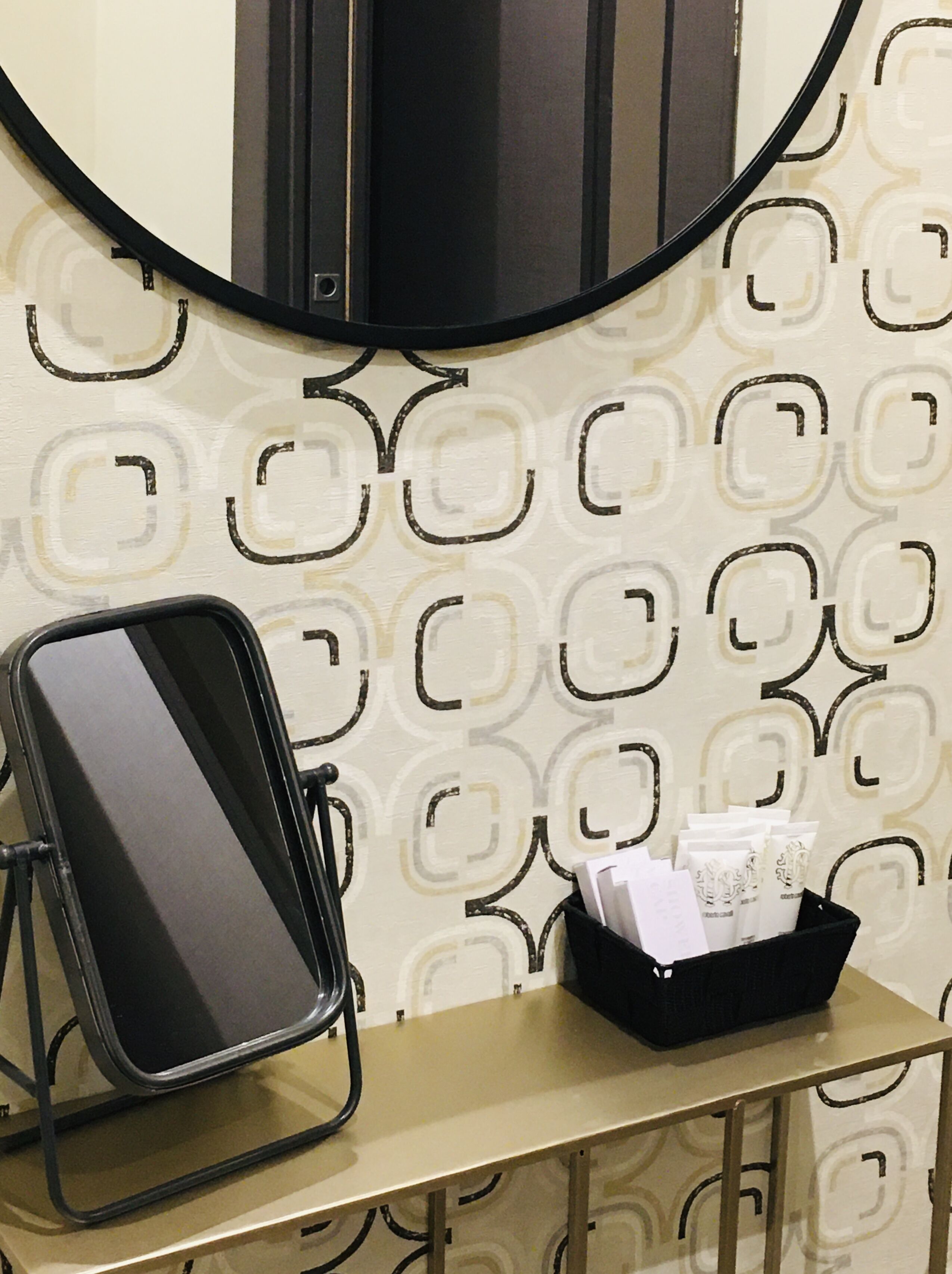 Una habitación con un espejo circular, una silla negra, una mesita con un atril de artículos de aseo y papel tapiz estampado.