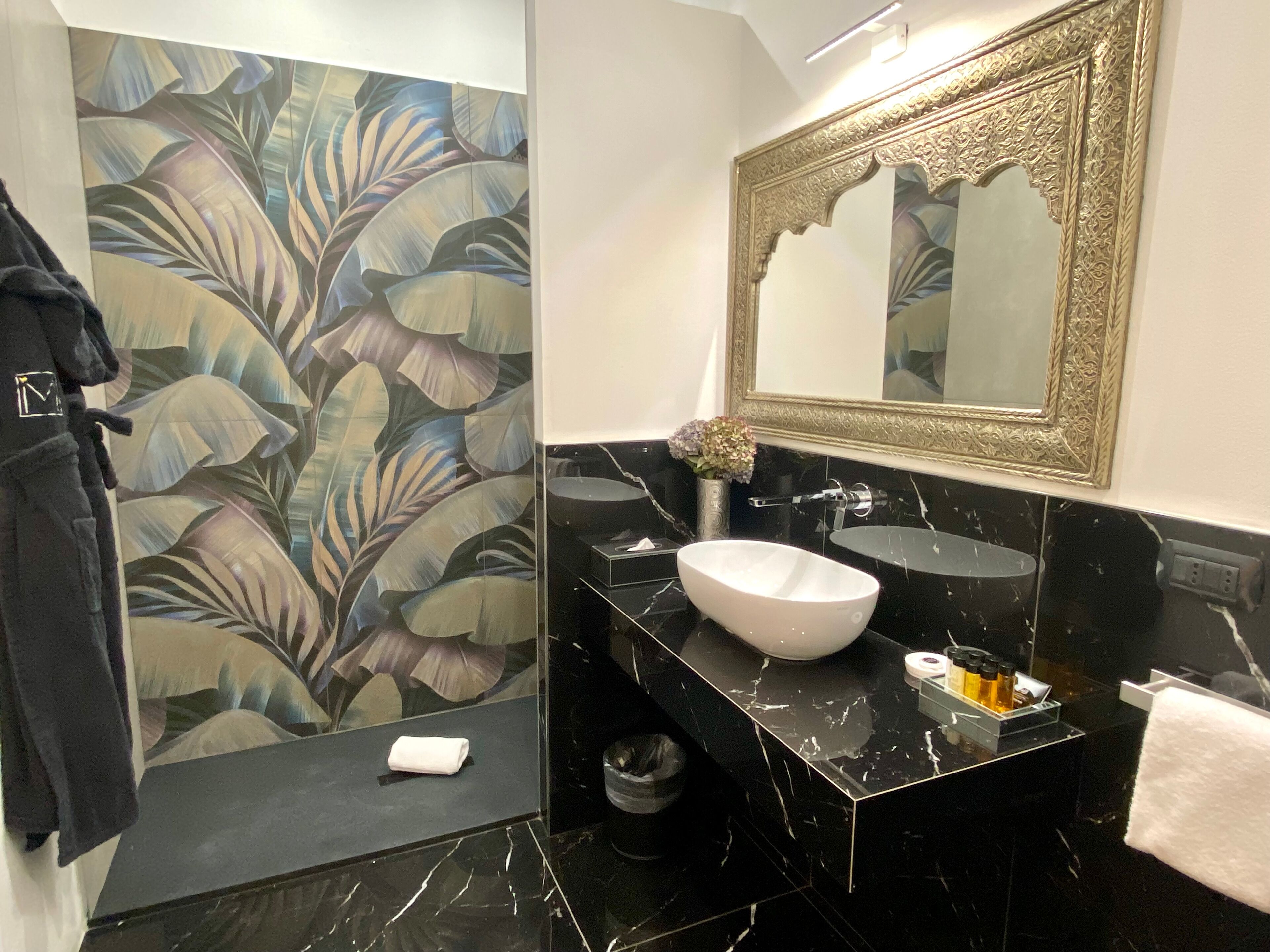 Ein Badezimmer mit einem schwarzen Marmorwaschbecken, einem großen Spiegel mit aufwendigem Rahmen und einem floralen Wanddesign.