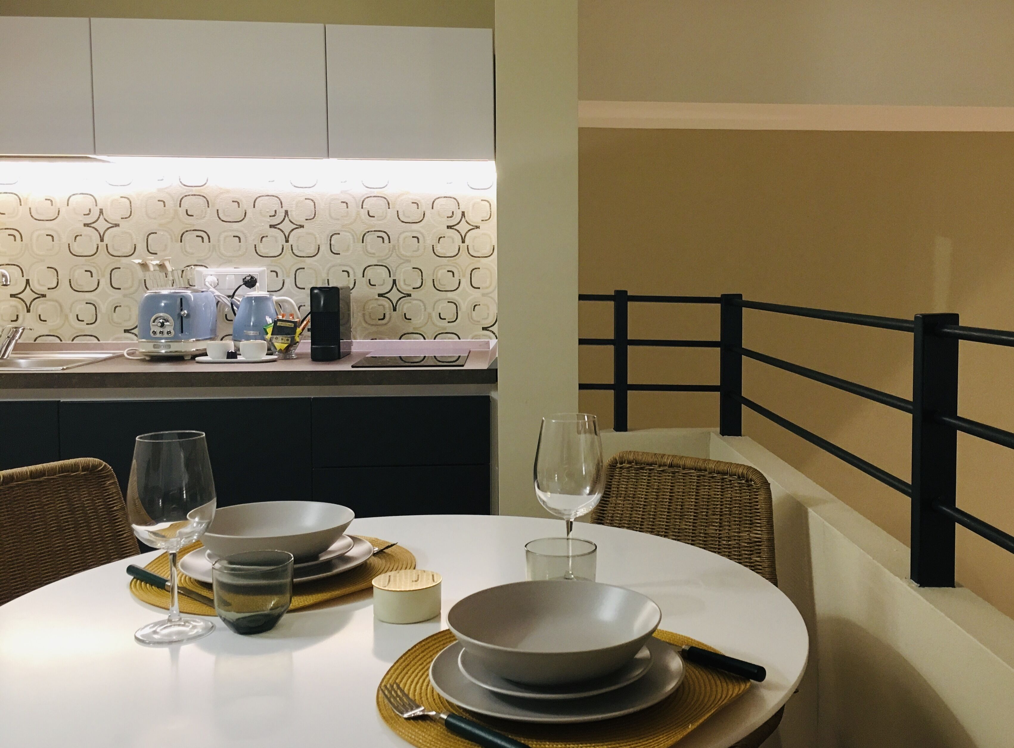 Una mesa de comedor dispuesta con platos, fuentes, cubiertos y copas de vino, con una cocina al fondo.