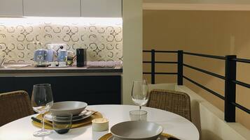 Una mesa de comedor dispuesta con platos, fuentes, cubiertos y copas de vino, con una cocina al fondo.