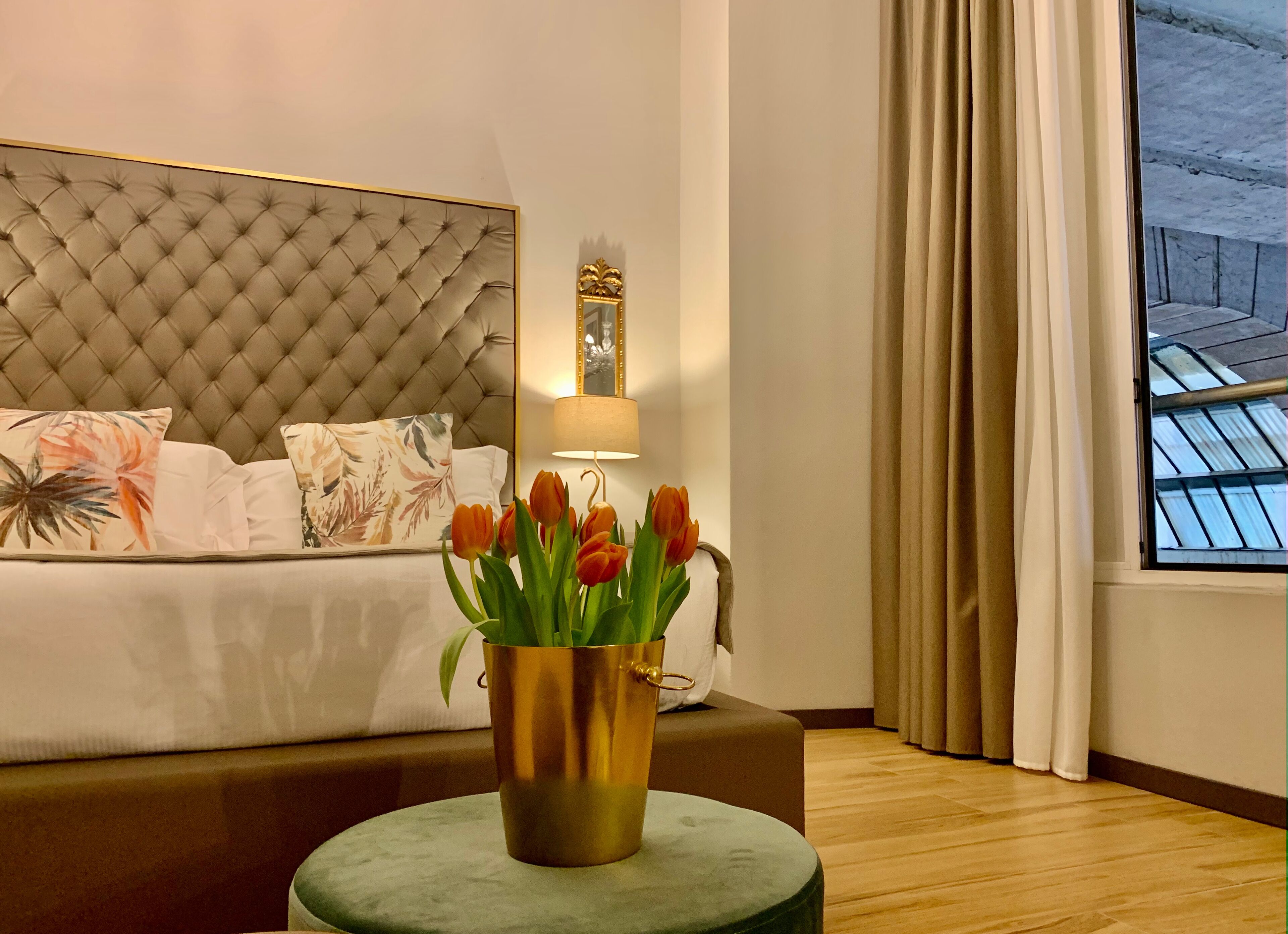 Un dormitorio con cabecera capitonada, cama con almohadas florales, lámpara de noche, florero con tulipanes y ventana con cortinas.